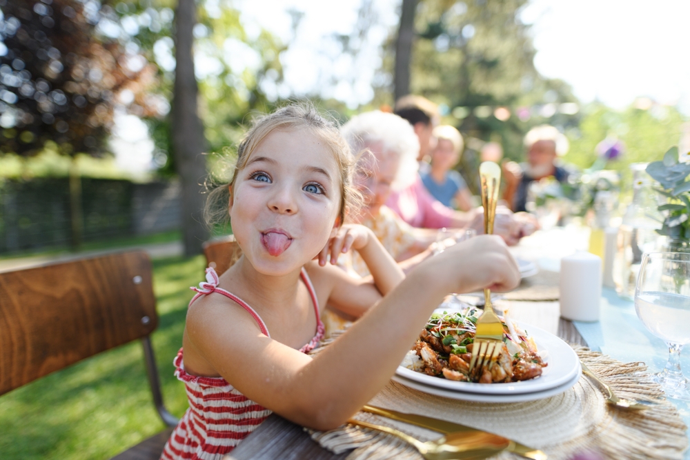 A little girl poking her tongue out while sitting at the backyard BBQ table.