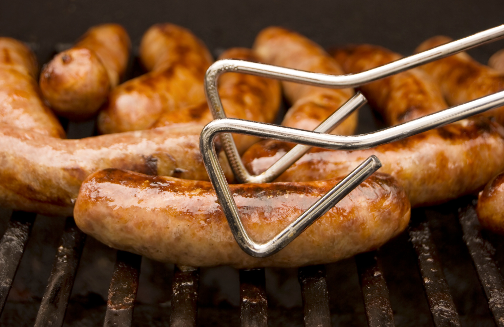 A pair of tongs picking up a perfectly cooked sausage on a BBQ grill.