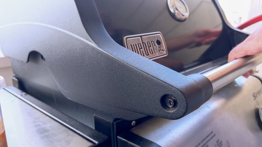 A person's hand resting on the handle of the Weber BBQ lid about to open it.