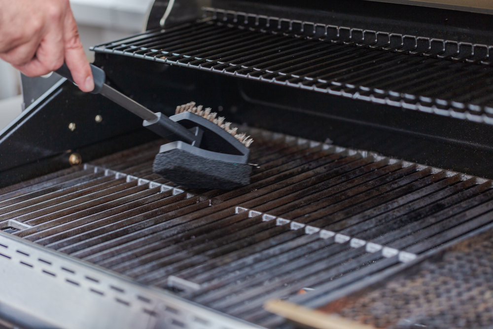 A person's hand holding a BBQ cleaning brushing cleaning the grill of a Weber BBQ.