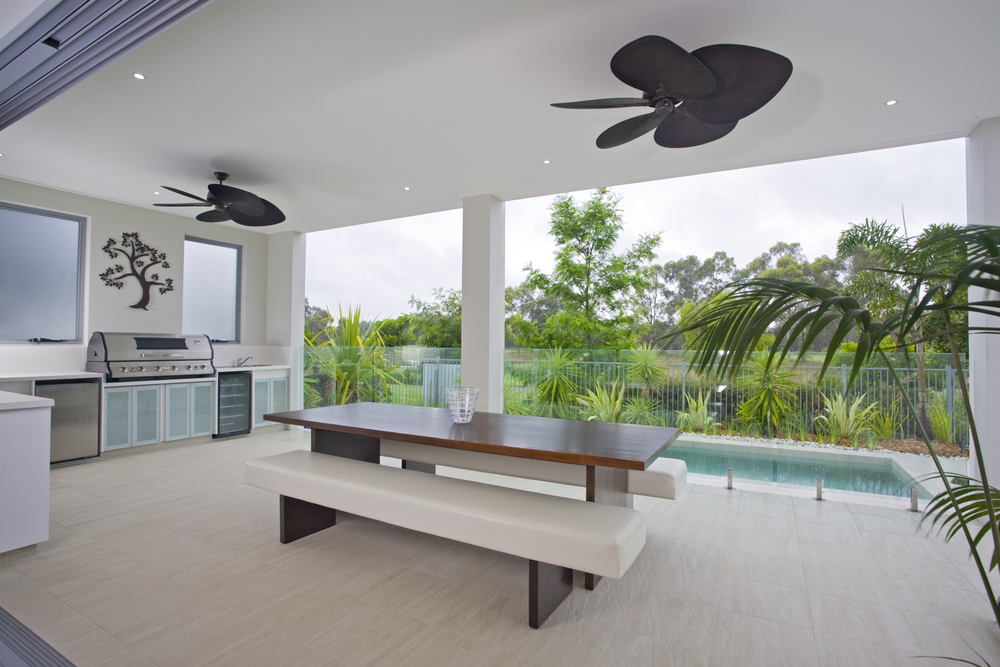 An outdoor kitchen area with a table, BBQ, pool and black ceiling fans. 