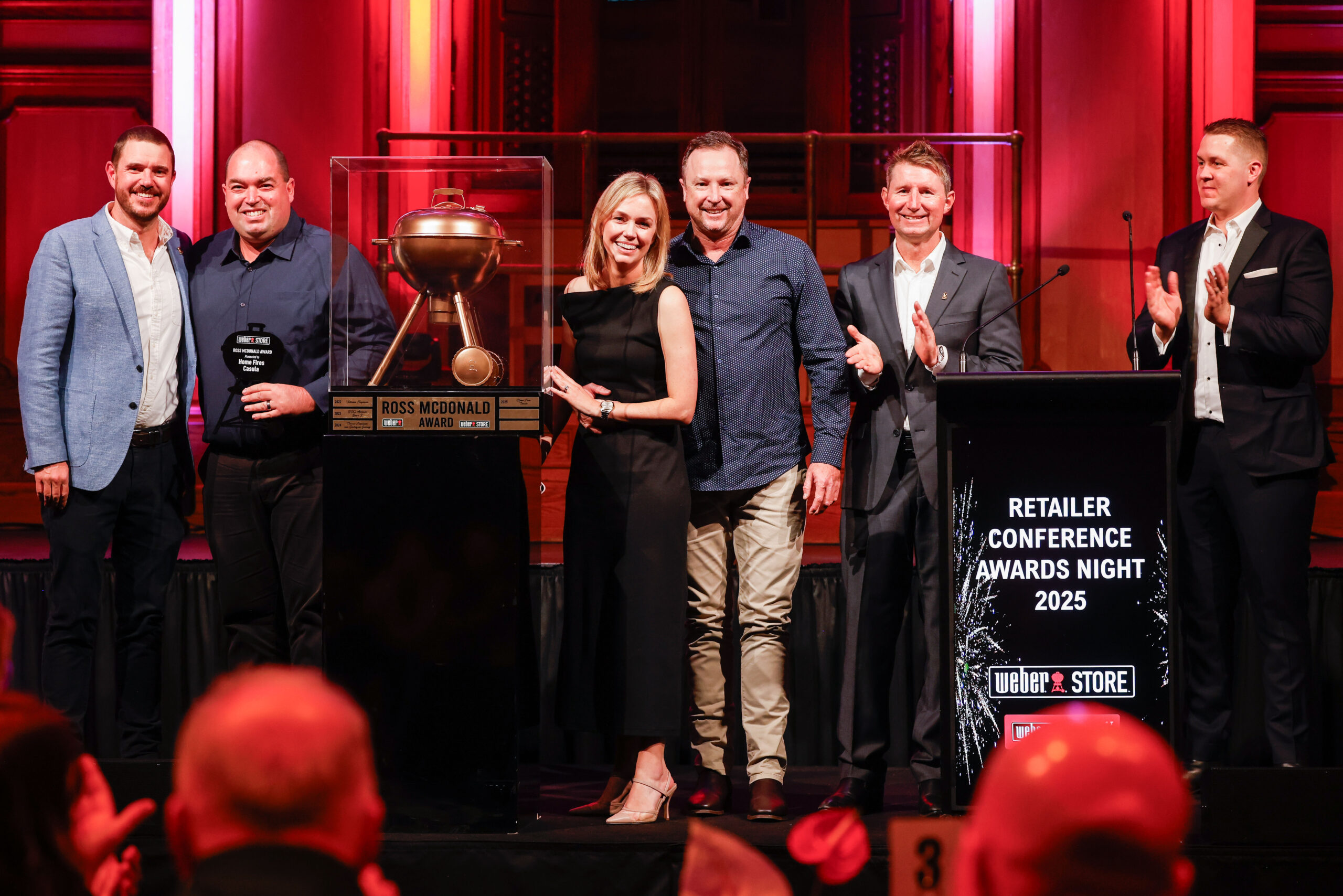 Home Fires accepting the Ross McDonald Award for Most Outstanding Weber Store 2025 for Australia and New Zealand at the Retailer Conference Awards Night. The award announcers are applauding and Home Fires team look very pleased.