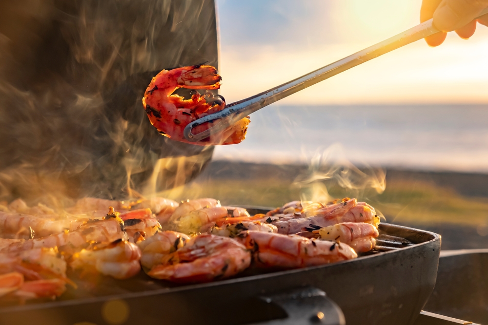 A person grilling prawns on a BBQ near the beach.