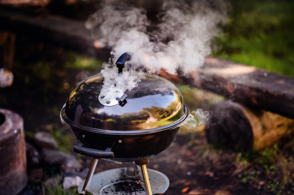 A Weber Kettle charcoal BBQ smoking in a forest near a campsite.
