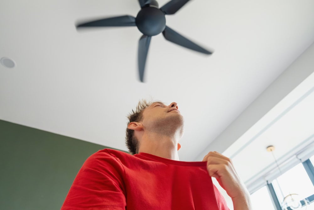 A man catching the breeze from below a ceiling fan. He is holding his red shirt open with relief on his face. 