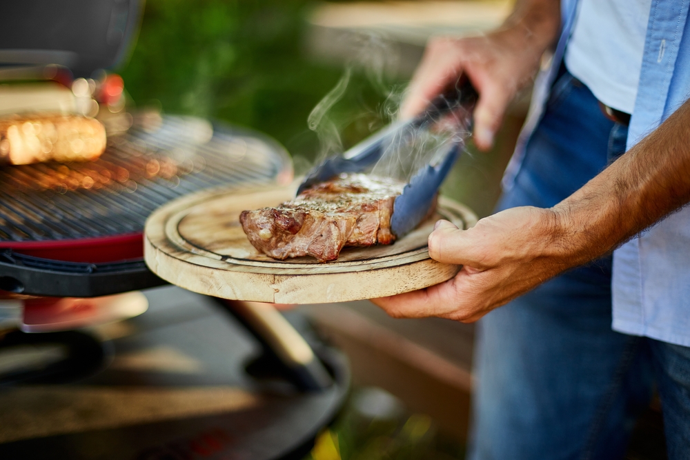 A man placing a barbecued steak onto a wooden platter board. There is a Weber BBQ in the background. 