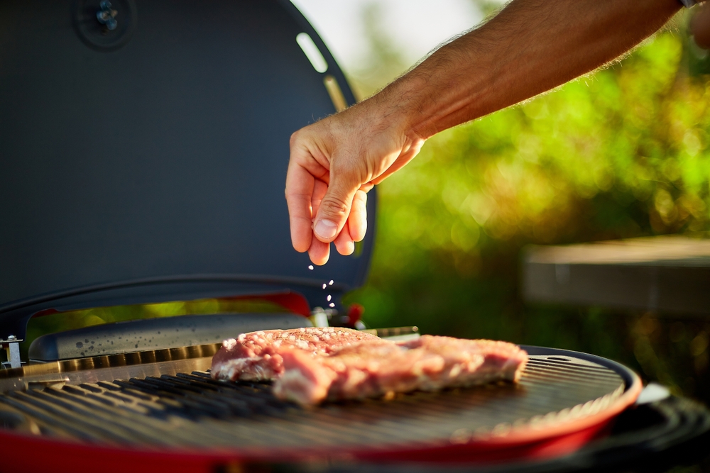 A man's hand sprinkling salt onto a steak grilling on a Weber Baby Q.
