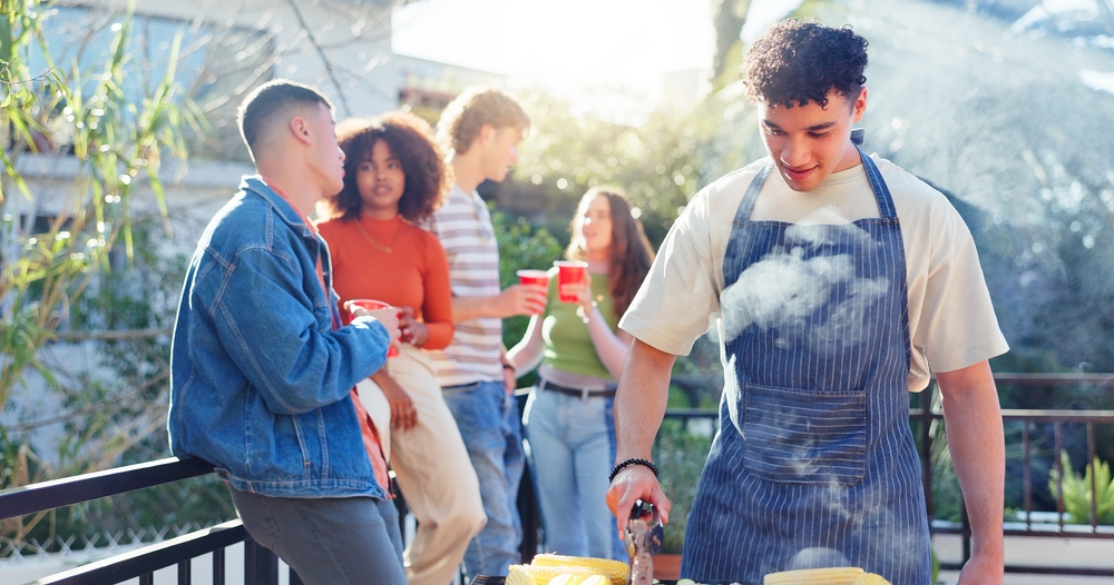 A group of friends using a Weber in Sydney during a sunny afternoon. There is one man cooking on the BBQ while four other friends stand with casually chatting.