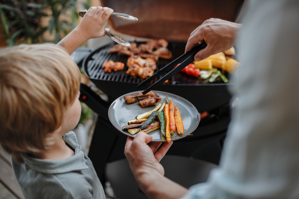 A man serving food onto a plate from a Weber Baby Q while a child helps.