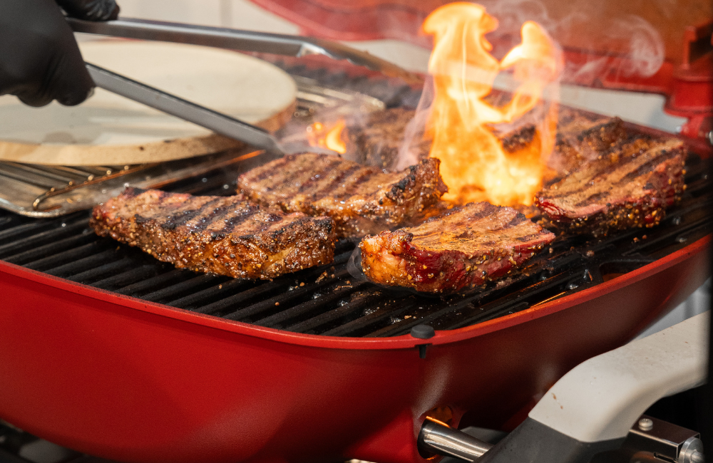 Cuts of meat grilling on a Weber baby Q with flames licking the edges.