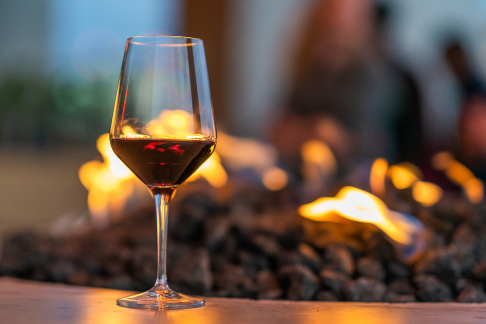 A wine glass with some red wine in it by an outdoor ethanol fire pit. 