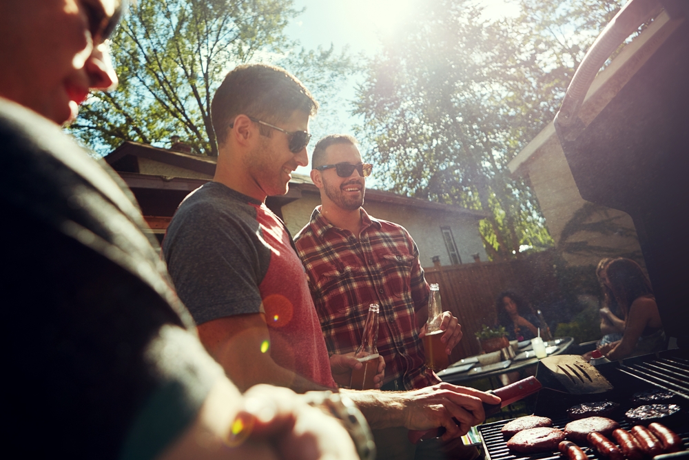 Three men standing around a BBQ while others sit at the table discussing the best BBQ for summer in Australia. 