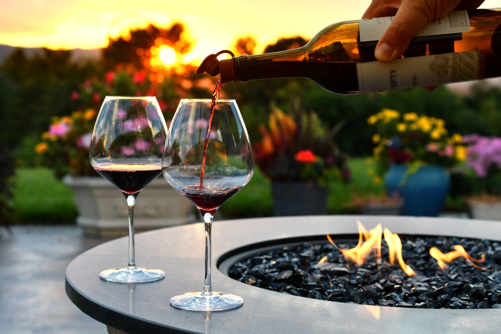 Two wine glasses on the edge of an ethanol firepit with a bottle of red win being poured.