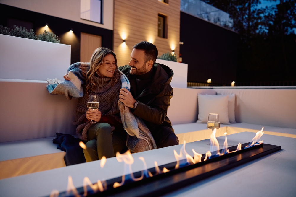 A couple sitting by an ethanol fire pit enjoying a wine while the male wraps the female in a blanket. 
