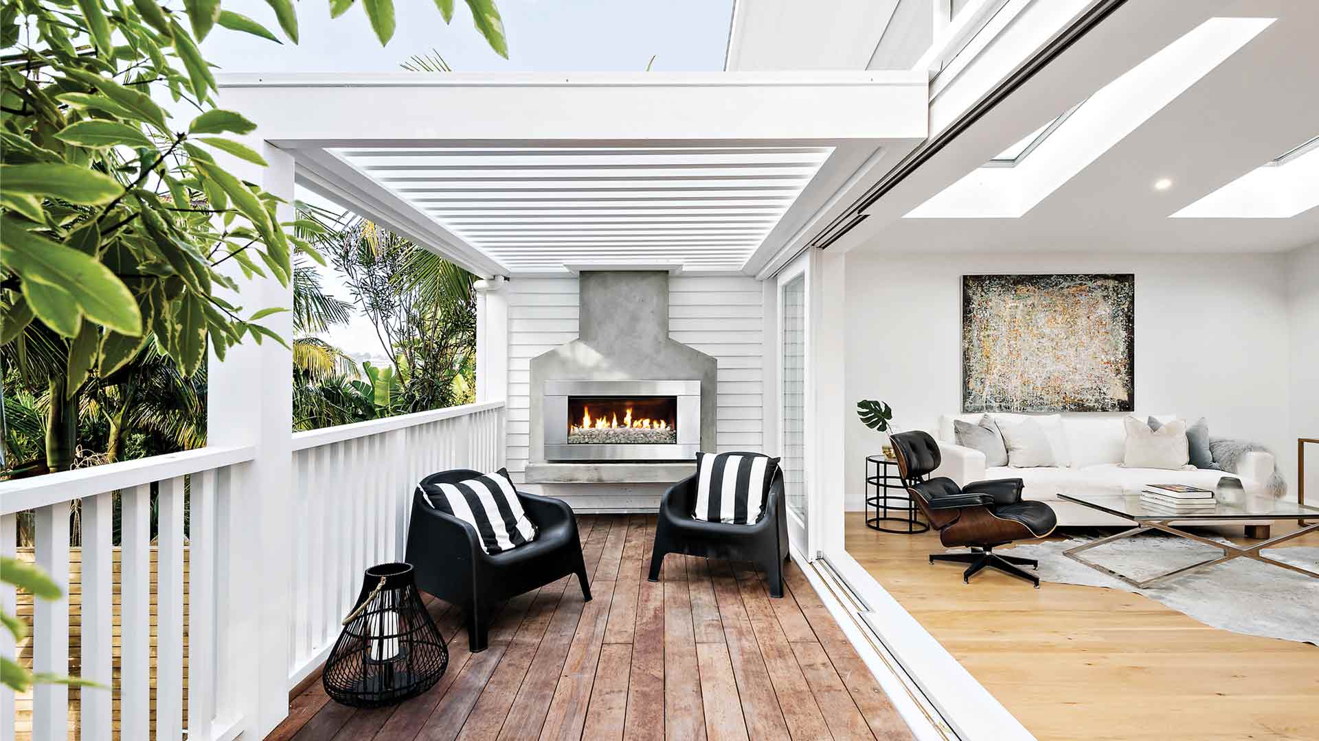 A modern built-in outdoor gas fireplace with a pebble fuel bed seamlessly connecting a covered timber deck to an indoor living room.