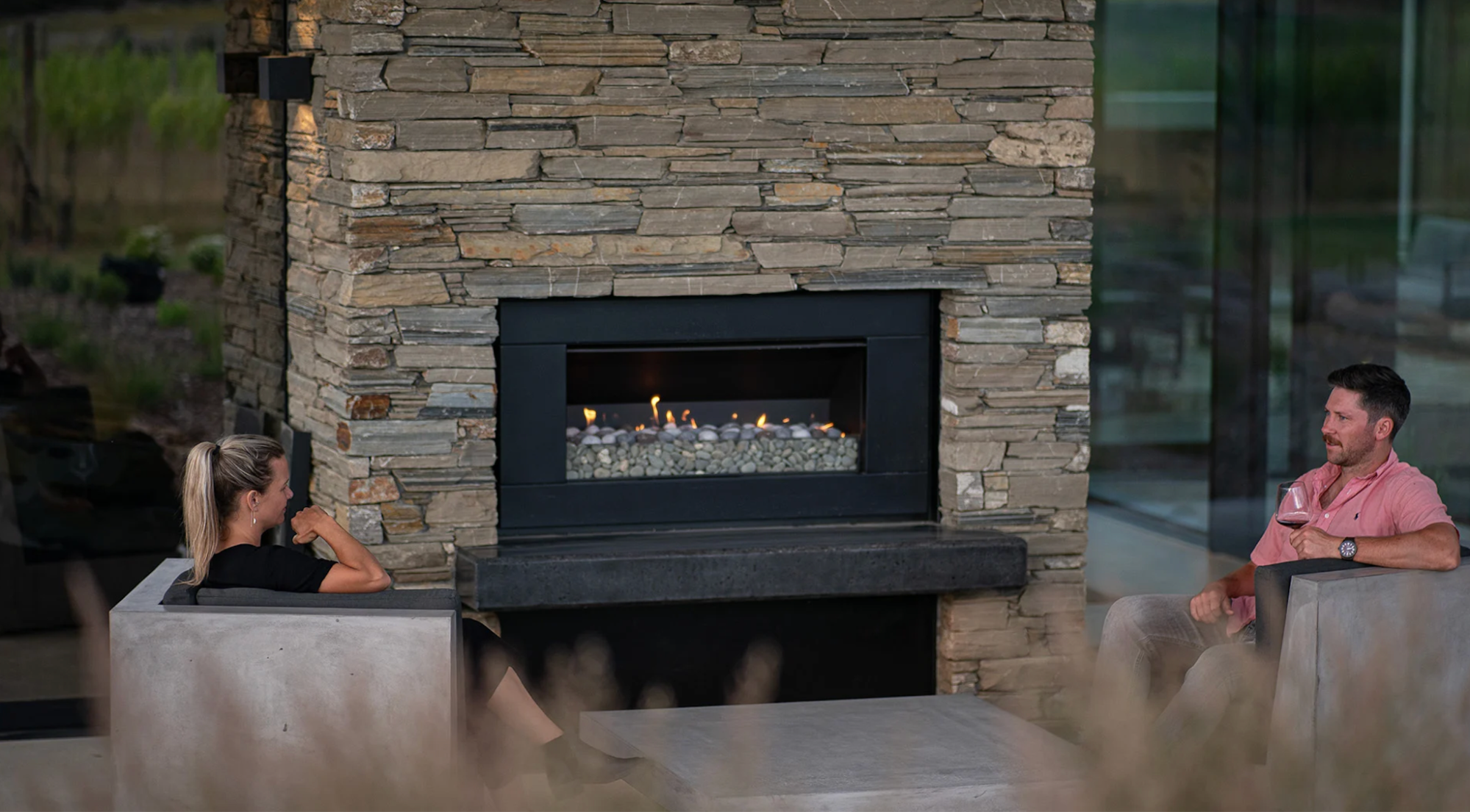 An Escea EF5000 outdoor gas fireplace built into a rustic stone feature wall, providing a cozy atmosphere for two people relaxing in an open-air lounge space.