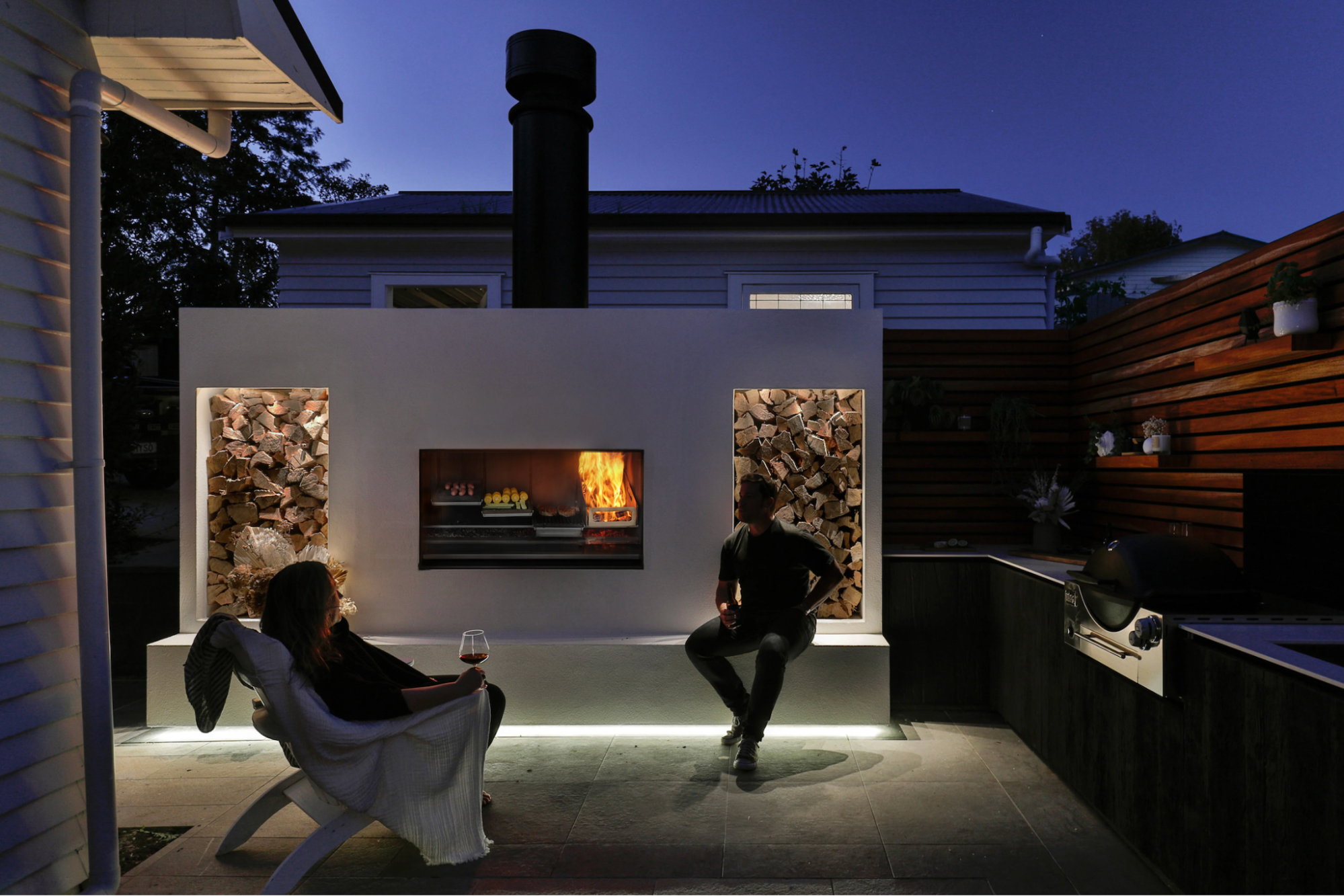 An Escea EK1250 outdoor wood fireplace kitchen lighting up a backyard entertaining area and integrated barbecue deck at dusk.