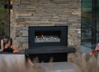An Escea EF5000 outdoor gas fireplace built into a rustic stone feature wall, providing a cozy atmosphere for two people relaxing in an open-air lounge space.
