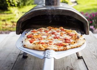 A delicious-looking pizza is being pulled out of a multi-fuel pizza oven in a backyard.